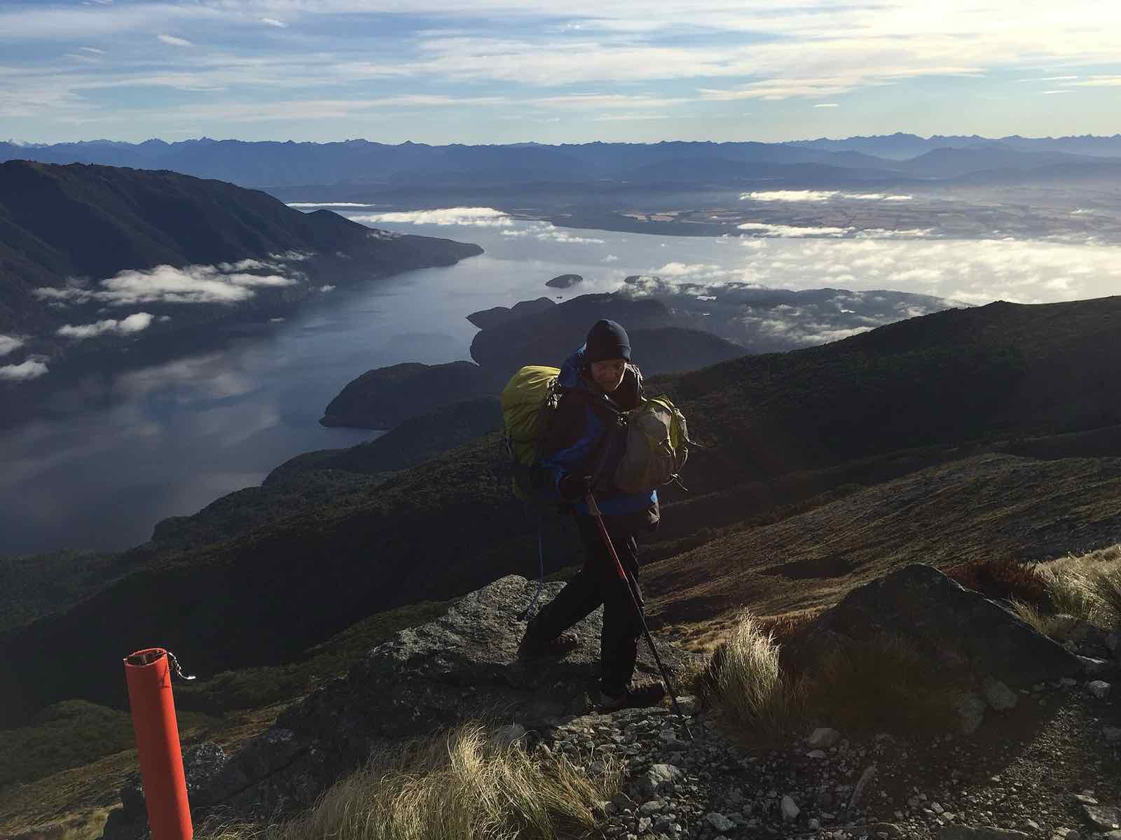 A tramper with a pack on a rocky slope high above Lake Te Anau and low cloud