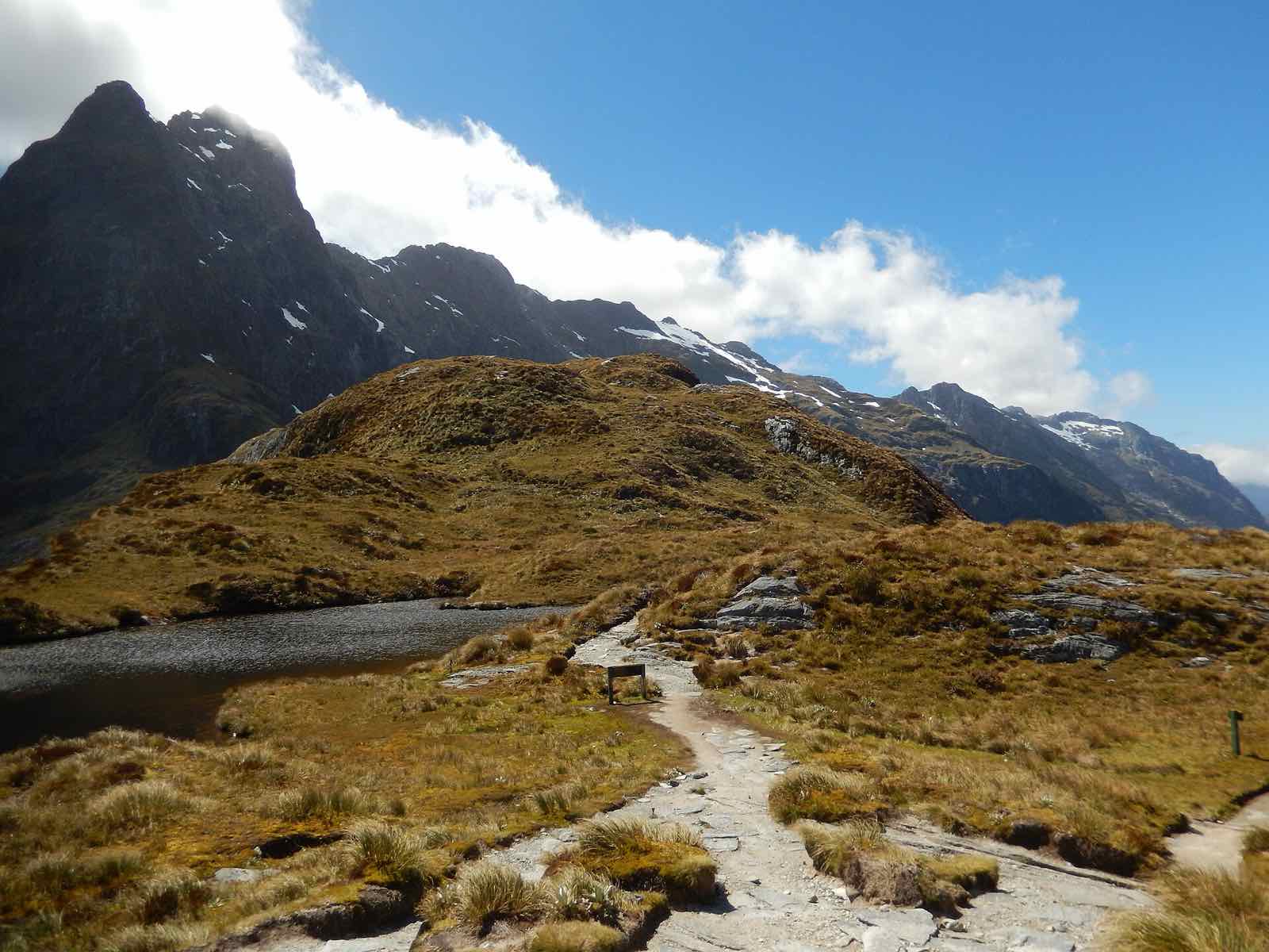 A gravel path crossing golden tussock beside a tarn near the top of McKinnon Pass, with peaks behind