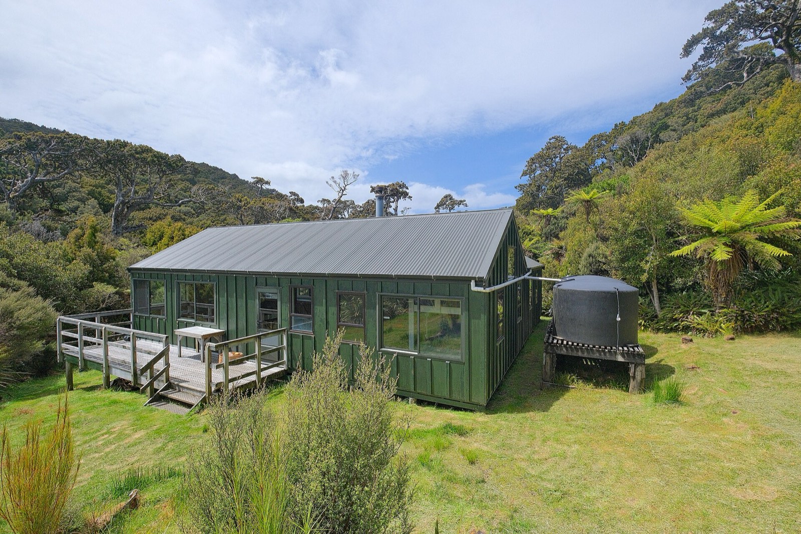 A green wooden hut with a deck and a water tank on a grassy clearing in the bush
