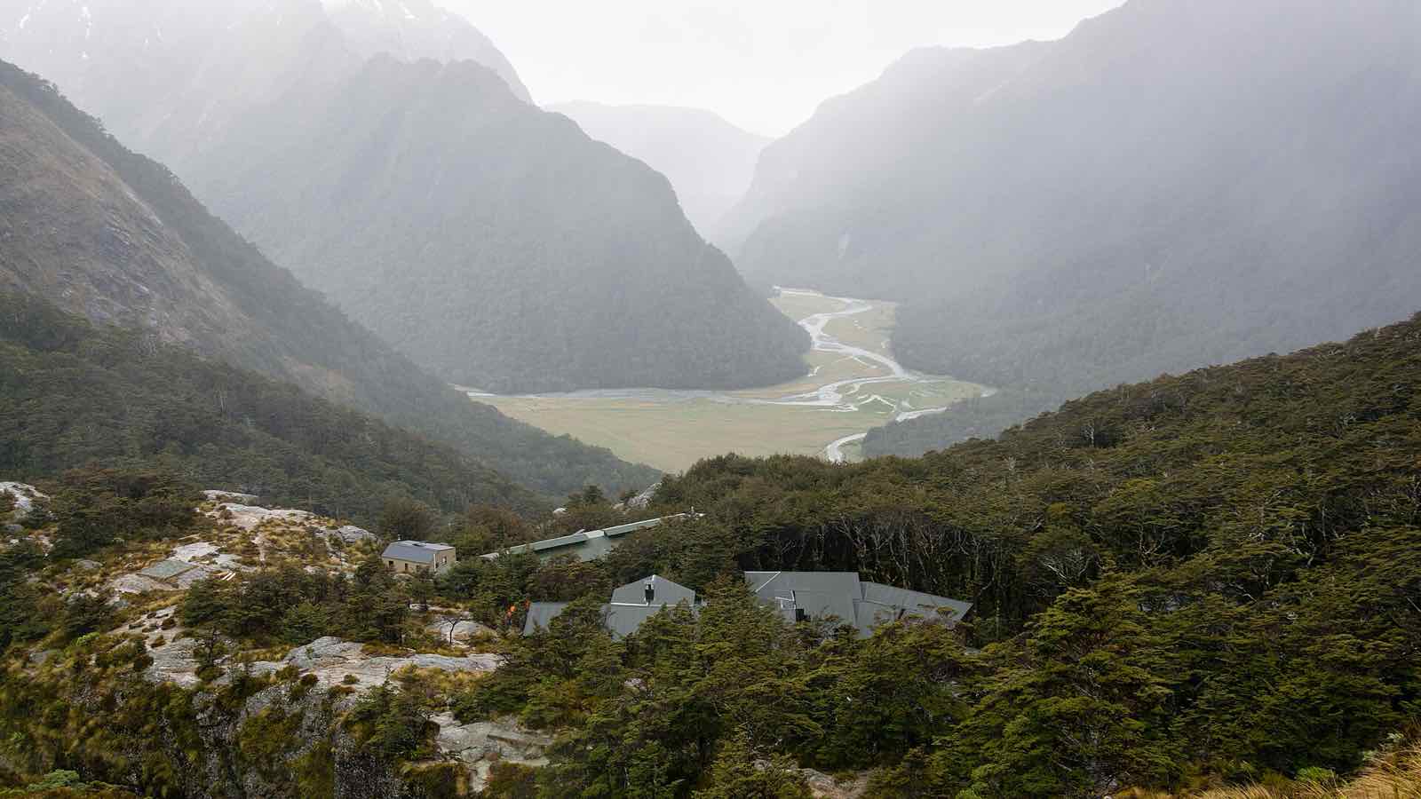 Hut buildings on a slope above a wide valley floor with a braided river