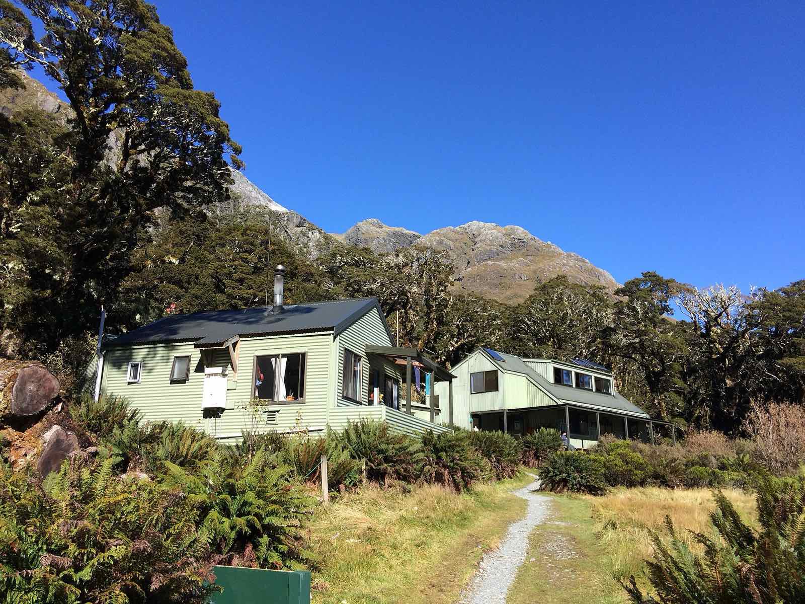 A pale green wooden hut in a sunny clearing with mountains behind
