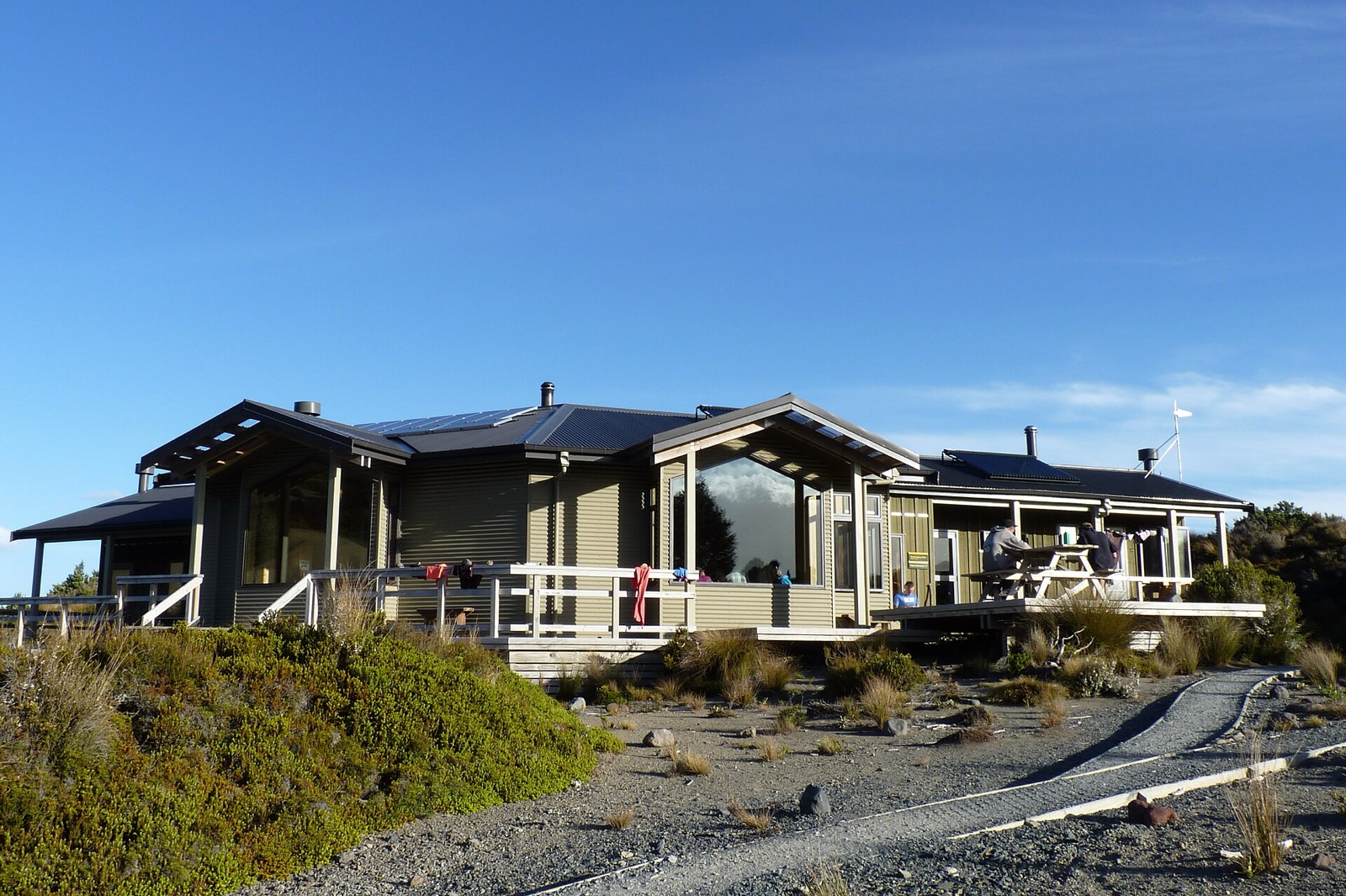 A modern timber hut with a wide deck and solar panels, with people at a picnic table outside