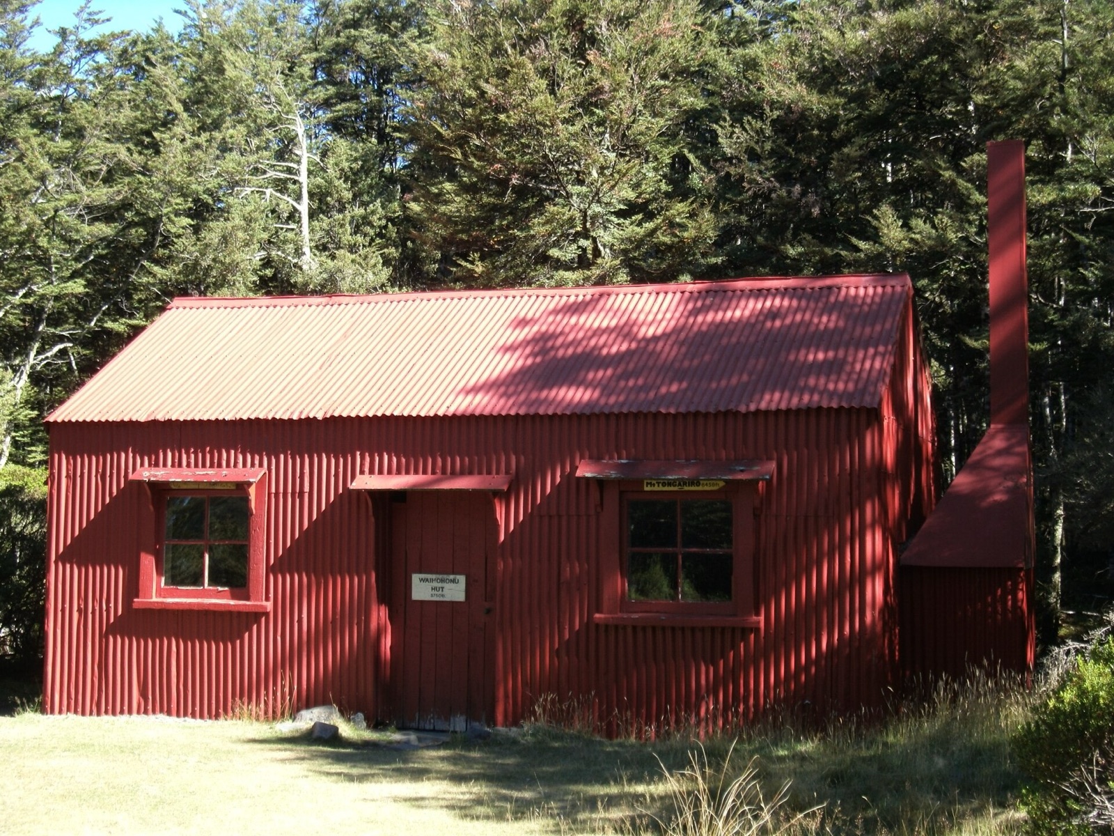A small red corrugated-iron hut with a tall chimney at the edge of beech forest