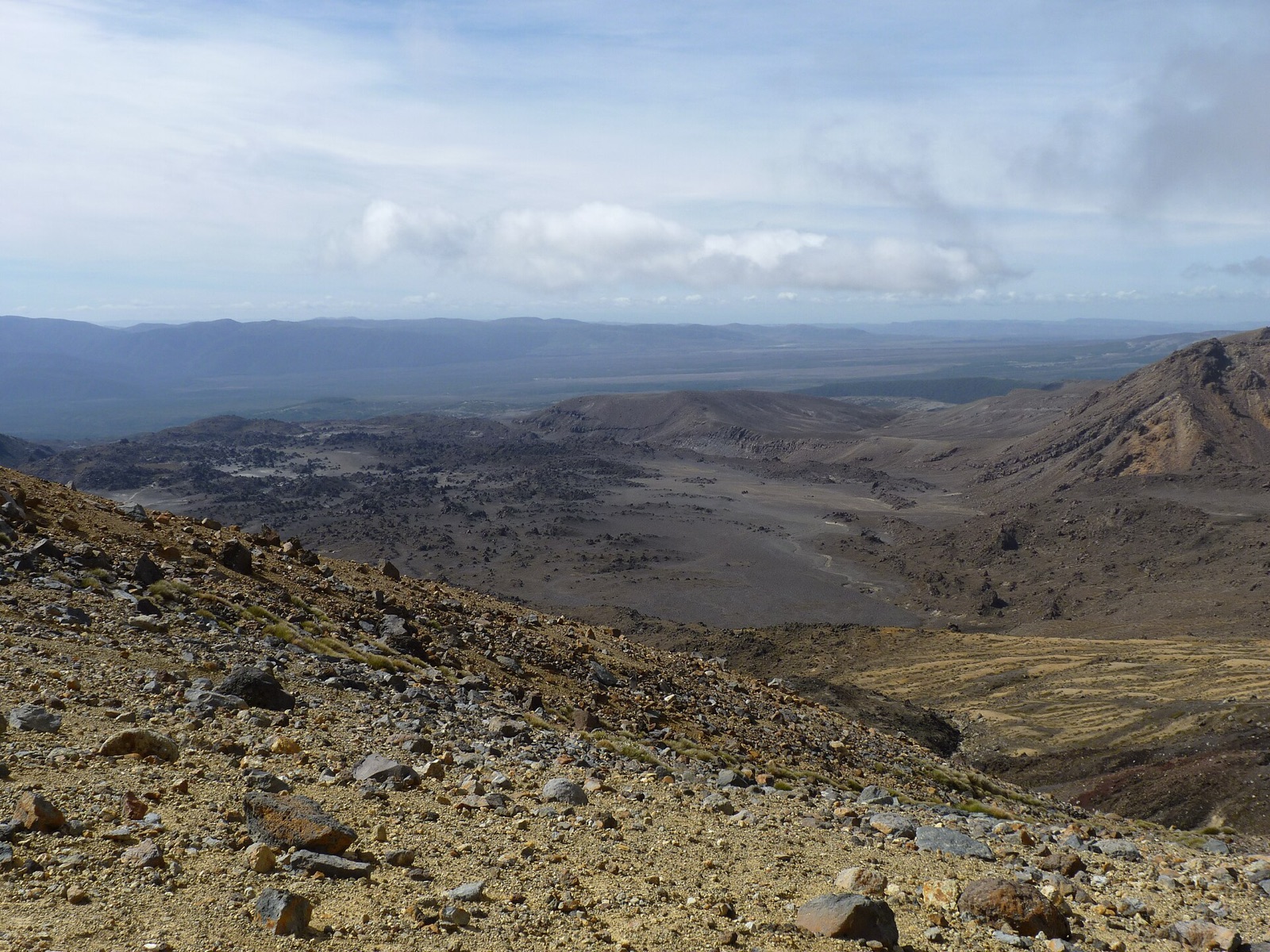 A rocky slope dropping to a wide valley floor of lava flows, with ranges in the distance