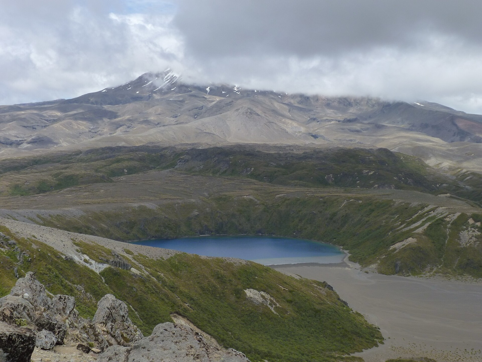 A blue crater lake in a tussock basin, with the slopes of Mount Ruapehu rising into cloud behind